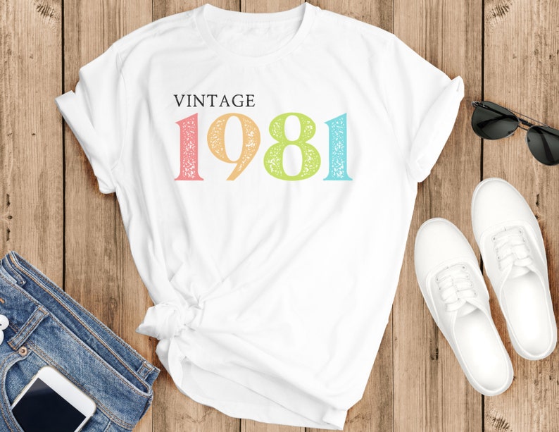 Puede incluir: Camiseta blanca con el texto "Vintage 1981" en colores arco&iacute;ris. Los n&uacute;meros est&aacute;n desgastados y tienen un aspecto vintage.