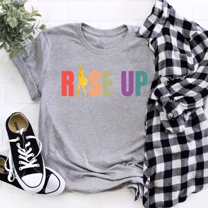 May include: A gray t-shirt with the word "RISE UP" printed in rainbow colors. The letters are outlined in black and the word "RISE" has a silhouette of a person dancing in the middle of the word.