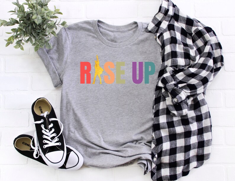 May include: A gray t-shirt with the word "RISE UP" printed in rainbow colors. The letters are outlined in black and the word "RISE" has a silhouette of a person dancing in the middle of the word.