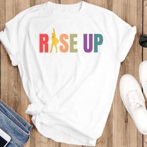 May include: White t-shirt with a rainbow colored graphic that says "RISE UP" and a yellow silhouette of a person raising their fist in the air.