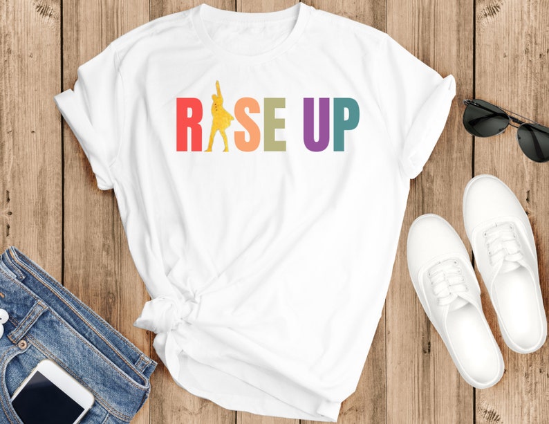 May include: White t-shirt with a rainbow colored graphic that says "RISE UP" and a yellow silhouette of a person raising their fist in the air.