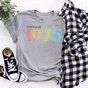 May include: Gray t-shirt with the text "Vintage 1975" in rainbow colors. The numbers are in a distressed font.