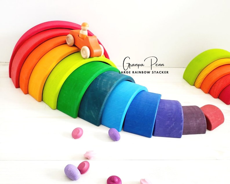 May include: A large rainbow wooden arch toy with a small wooden car rolling down the top of the arch. The toy is made of natural wood and is painted in bright rainbow colors. The toy is called "Grandpa Penn Large Rainbow Stacker".
