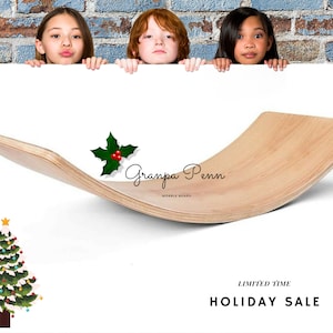 Puede incluir: Una tabla oscilante de madera de color claro con un dise&ntilde;o curvo. El texto "Granpa Penn Wobble Board" es visible. Tambi&eacute;n hay un &aacute;rbol de Navidad y una ramita de acebo. La imagen incluye a tres ni&ntilde;os asom&aacute;ndose por encima de una superficie blanca.