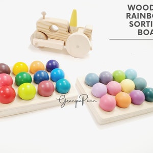 Puede incluir: Tablero de clasificación de arcoíris de madera con esferas de colores. El tablero presenta un juguete de tractor y dos bandejas de madera con esferas pintadas en varios colores, incluyendo rojo, amarillo y azul. El texto "WOODEN RAINBOW SORTING BOARD" es visible.
