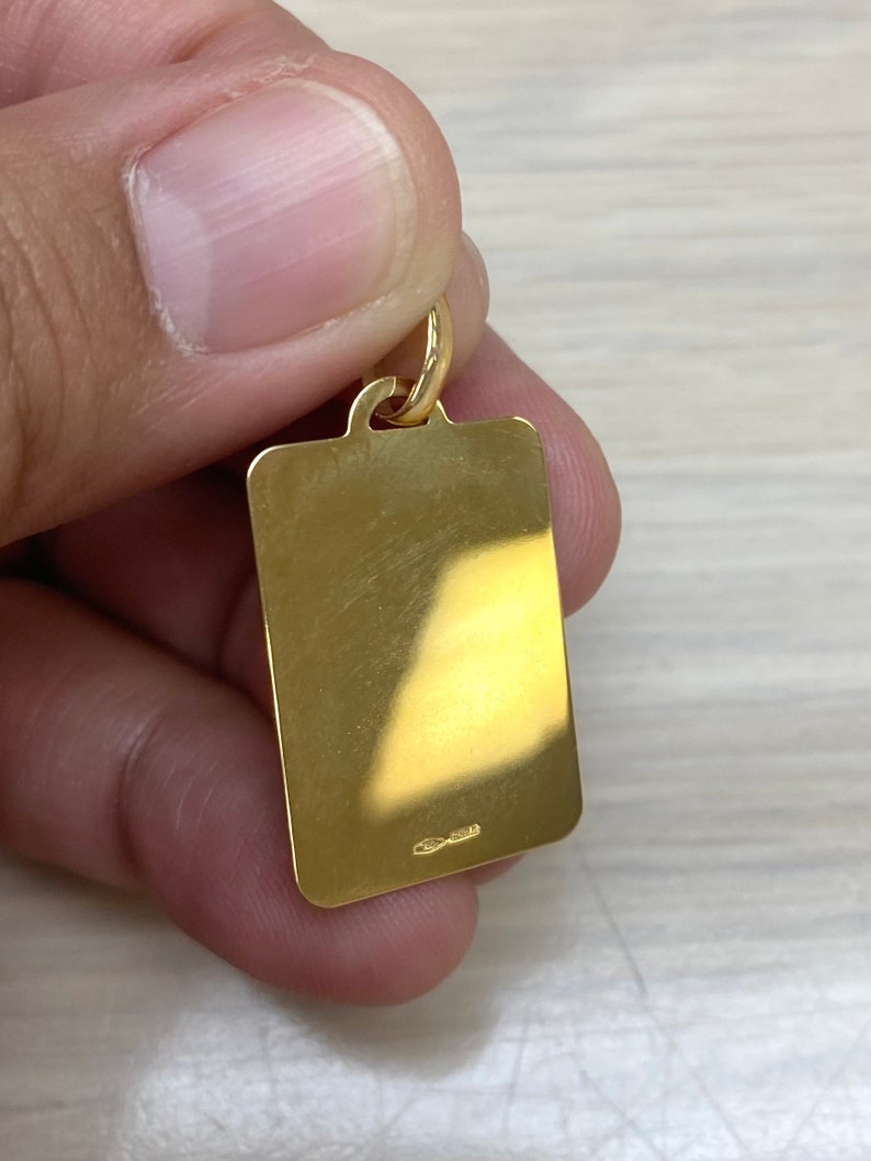 Pode incluir: Um pingente retangular dourado com uma superf&iacute;cie lisa e um pequeno la&ccedil;o na parte superior. O pingente tem uma pequena inscri&ccedil;&atilde;o na parte inferior.