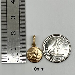 May include: A gold-colored pendant featuring a cherub design. The pendant is 10mm in diameter and is shown next to a Canadian 10-cent coin for size comparison.
