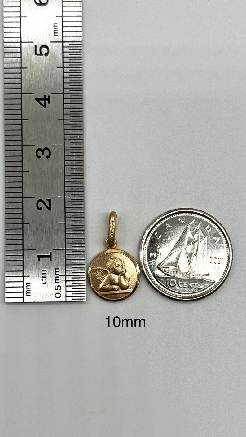 May include: A gold-colored pendant featuring a cherub design. The pendant is 10mm in diameter and is shown next to a Canadian 10-cent coin for size comparison.