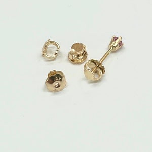 Op de afbeelding: Een verzameling gouden oorbellen en oorbelruggen. Eén oorbel heeft een kleine roze edelsteen. De oorbelruggen omvatten vlindersluitingen en schroefmodellen. De sieraden worden gepresenteerd op een wit oppervlak.