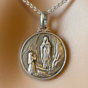 May include: Silver chain necklace with a round pendant featuring a depiction of the Virgin Mary and a kneeling woman.