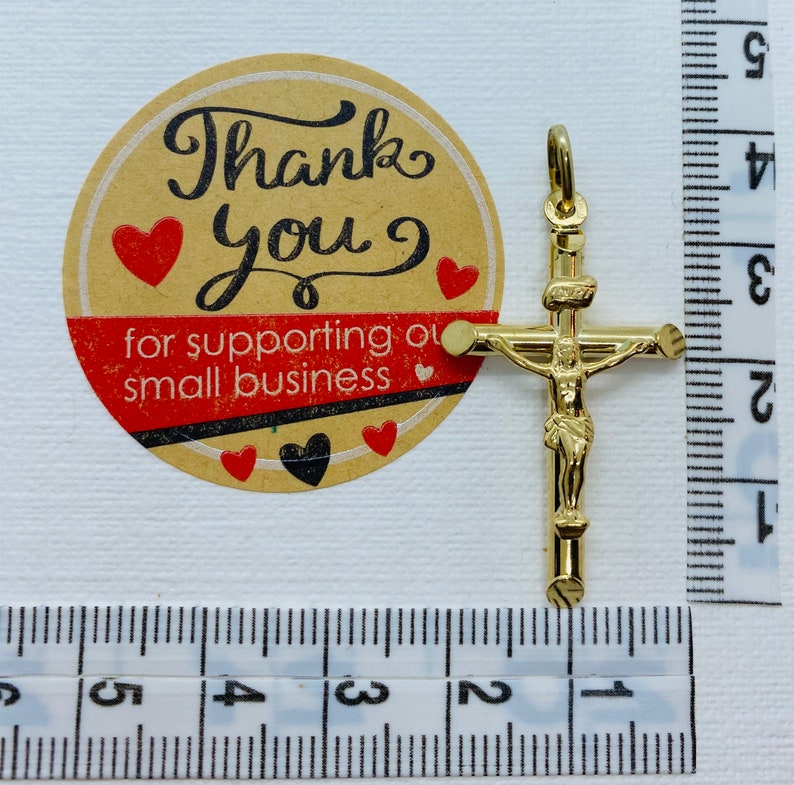 May include: Gold-toned crucifix pendant with a figure of Jesus Christ on the cross. The pendant is approximately 2 cm in length and is shown next to a ruler for size reference.