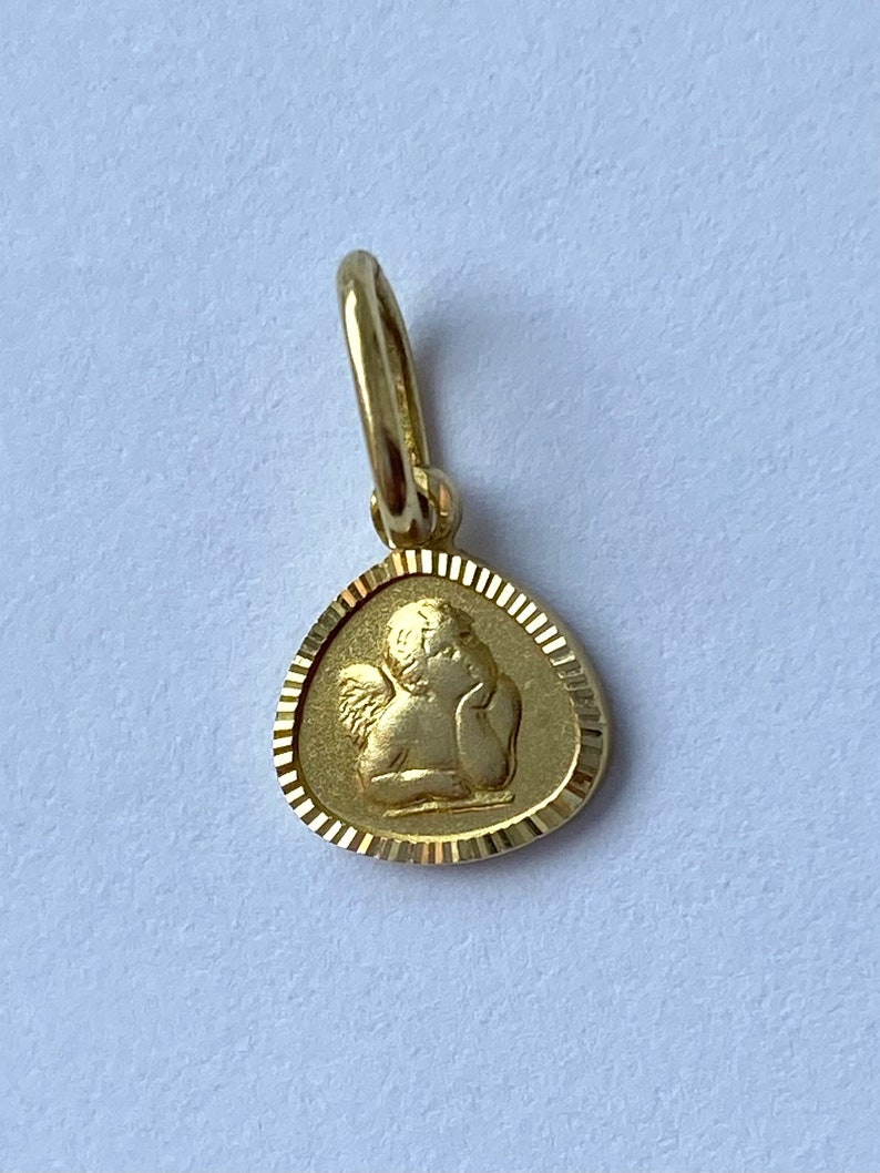 May include: A gold-colored pendant featuring a detailed image of a cherub angel with wings. The pendant is oval-shaped with a textured border.