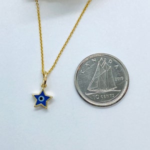 Może przedstawiać: Złoty łańcuszek z małym niebieskim i białym amuletem w kształcie gwiazdy z motywem złego oka. Amulet jest mniej więcej wielkości kanadyjskiej dziesięciocent&oacute;wki.