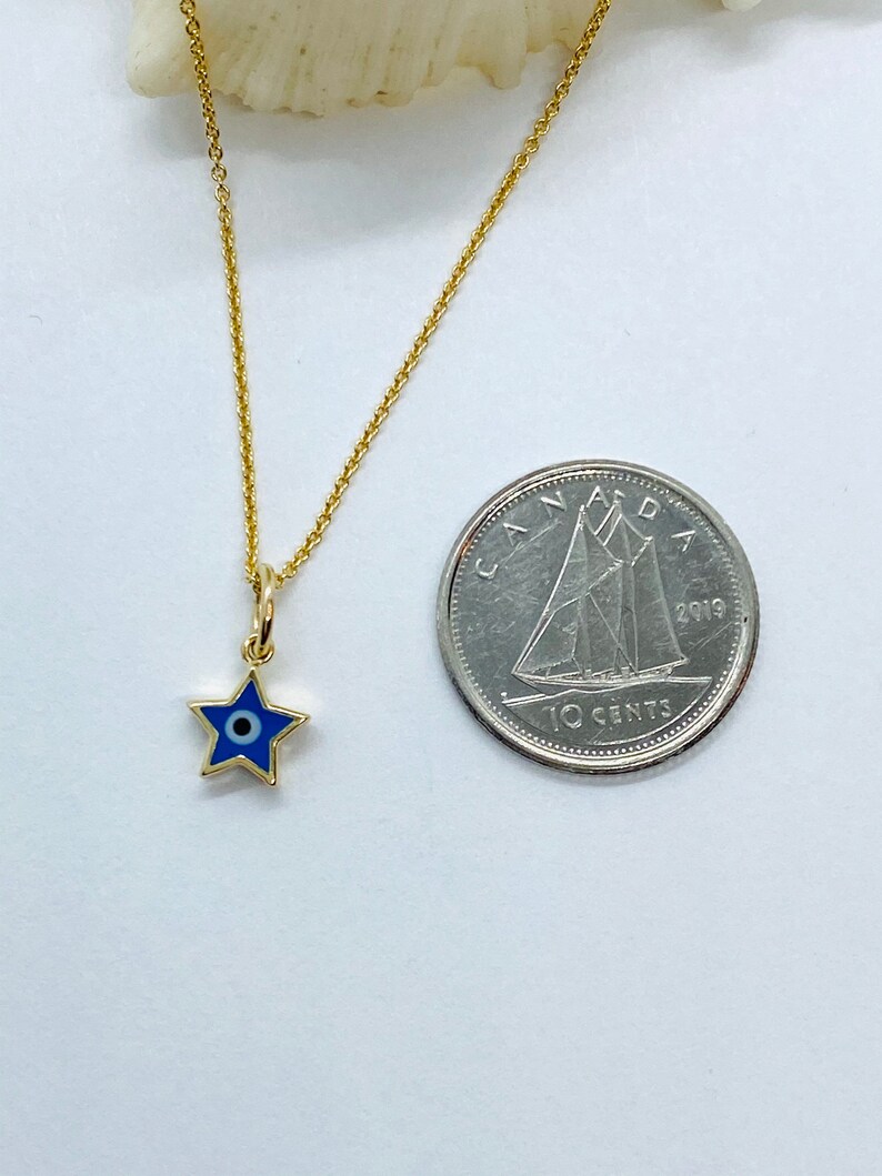 Może przedstawiać: Złoty łańcuszek z małym niebieskim i białym amuletem w kształcie gwiazdy z motywem złego oka. Amulet jest mniej więcej wielkości kanadyjskiej dziesięciocent&oacute;wki.