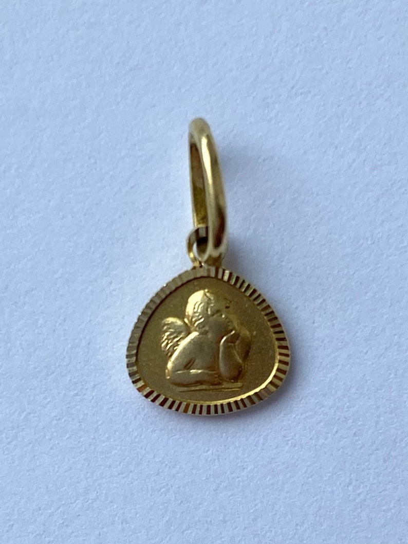 May include: A gold pendant featuring a detailed image of a cherub angel with wings. The pendant is shaped like a teardrop and has a textured border.