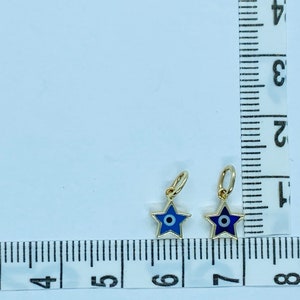 Może przedstawiać: Dwa złote charms w kształcie gwiazdy z niebieskimi wzorami złego oka. Charms mają około 1,25 cm i są pokazane na białym tle z linijką do skalowania.