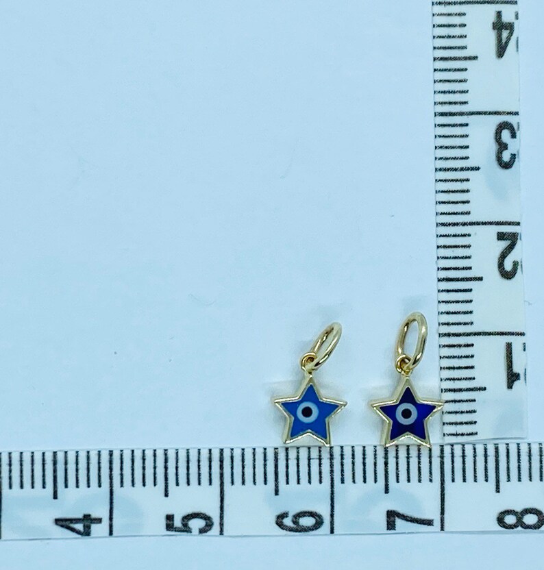 Może przedstawiać: Dwa złote charms w kształcie gwiazdy z niebieskimi wzorami złego oka. Charms mają około 1,25 cm i są pokazane na białym tle z linijką do skalowania.