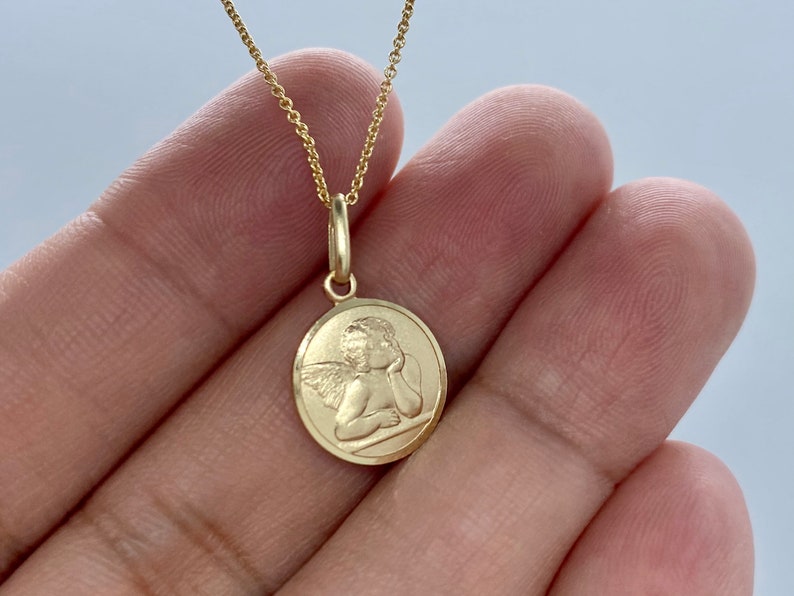 May include: A gold chain necklace with a round pendant featuring a detailed image of a cherub angel with wings.