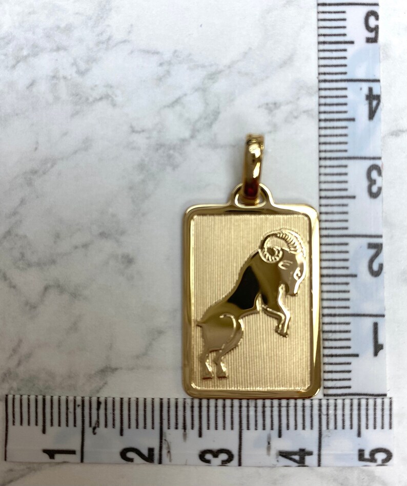 Pode incluir: Pingente dourado com uma representa&ccedil;&atilde;o detalhada do signo do zod&iacute;aco &Aacute;ries, um carneiro, dentro de uma moldura retangular. O pingente mede aproximadamente 2,5 cm de comprimento.