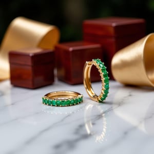 May include: A pair of gold hoop earrings with emerald green gemstones. The earrings are on a white marble surface.