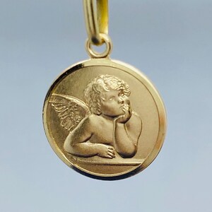 May include: A gold-colored pendant featuring a cherub with wings, resting its head on its hand. The cherub is depicted in a circular frame.