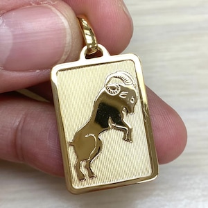 Pode incluir: Pingente dourado com uma representa&ccedil;&atilde;o detalhada do signo do zod&iacute;aco &Aacute;ries, o carneiro, dentro de uma moldura retangular.