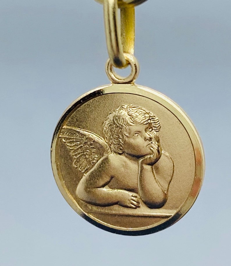 May include: A gold-colored pendant featuring a cherub with wings, resting its head on its hand. The cherub is depicted in a thoughtful pose, with a detailed face and intricate wings.