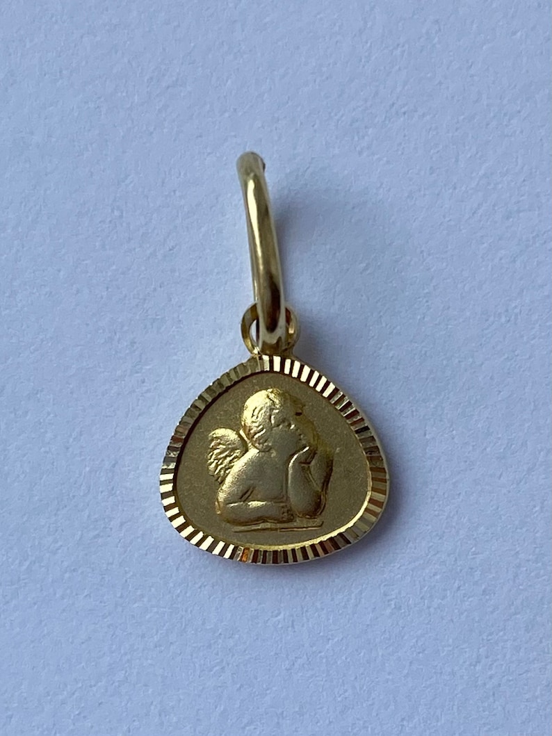 May include: A gold charm featuring a detailed depiction of a cherub angel with wings, resting its head on its hand. The charm is in the shape of a teardrop with a decorative border.