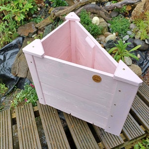 May include: A pink triangular wooden planter with a small wooden label on the front. The planter is sitting on a wooden deck.