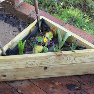 May include: A triangular wooden planter box with a variety of plants growing inside. The planter is made of light brown wood and has a natural finish. The plants include green leaves, yellow flowers, and purple flowers.