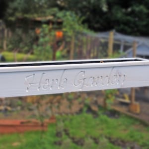 May include: White wooden planter box with the words "Herb Garden" carved into the front. The planter is hanging from a hook.