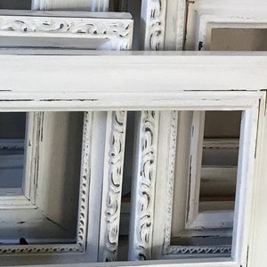 May include: A collection of white, ornate picture frames with distressed edges. The frames vary in size and design, some featuring detailed carvings and others with simpler, clean lines. The overall aesthetic is vintage and shabby chic.