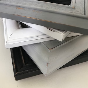 May include: A stack of four wooden picture frames in various colors: gray, white, light gray, and black. The frames have a distressed, rustic finish with visible wood grain and slight chipping, suggesting a vintage aesthetic.