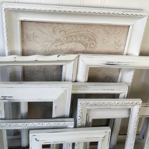 May include: A collection of distressed white picture frames in various sizes and styles. Some frames have ornate detailing, while others are more simple. The frames are arranged in a layered composition, with a patterned background visible in some.
