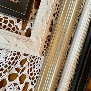 May include: An assortment of decorative picture frames in various colors and styles. The frames include white, black, and metallic gold finishes. Some frames have ornate detailing, while others have a simpler, more modern design. The frames are arranged on a white lace doily.