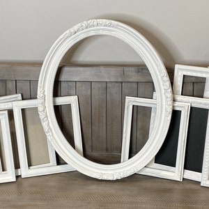 May include: A collection of white picture frames in various sizes and shapes, including rectangular and oval designs. Some frames have ornate detailing, while others are simpler. The frames are arranged on a wooden surface, ready for displaying photos or artwork.