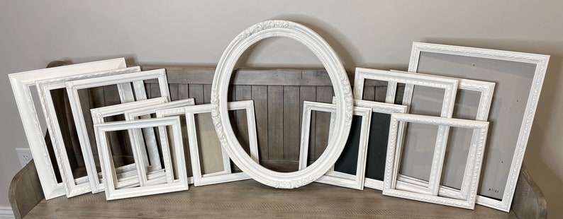 May include: A collection of white picture frames in various sizes and shapes, including rectangular and oval designs. Some frames have ornate detailing, while others are simpler. The frames are arranged on a wooden surface, ready for displaying photos or artwork.