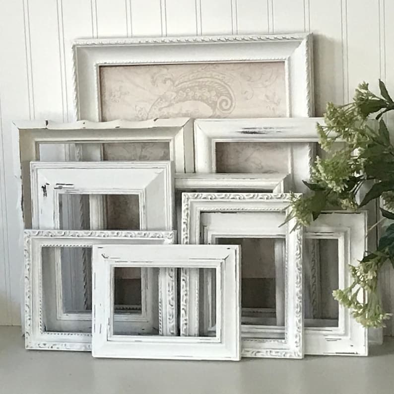 May include: A collection of distressed white picture frames in various sizes and shapes. Some frames have ornate detailing, while others are simpler. One frame contains a beige paisley print. The frames are arranged in a layered composition, suggesting a vintage or farmhouse aesthetic.