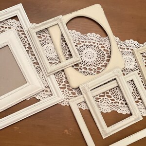 May include: A collection of white picture frames in various sizes and shapes, including rectangular and oval, arranged on a white lace doily. The frames have a distressed, vintage look, with visible texture and slight variations in color.