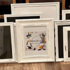 May include: A collection of white picture frames of various sizes and styles, including rectangular and square shapes. Some frames have ornate detailing, while others are more minimalist. One frame displays a print with text and botanical illustrations.