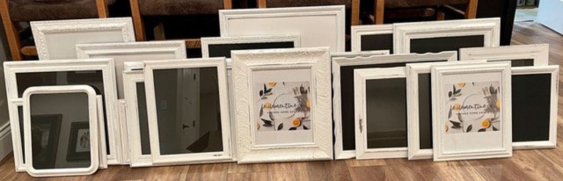 May include: A collection of white picture frames of various sizes and styles, including rectangular and square shapes. Some frames have ornate detailing, while others are more minimalist. One frame displays a print with text and botanical illustrations.