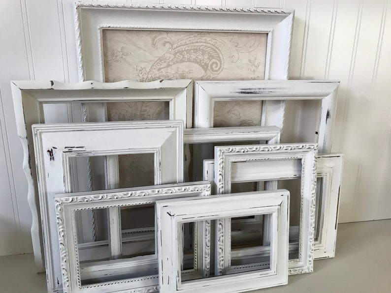 May include: A collection of white, distressed picture frames in various sizes and styles. Some frames have ornate detailing, while others have a simpler design. The frames are arranged in a layered composition, creating a vintage aesthetic.