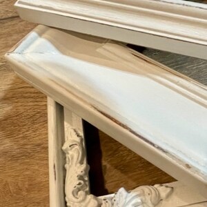 May include: Close-up of two white picture frames with ornate corner detailing. The frames have a distressed, antique finish with visible wood grain and a light blue undertone. The frames are stacked on top of each other.