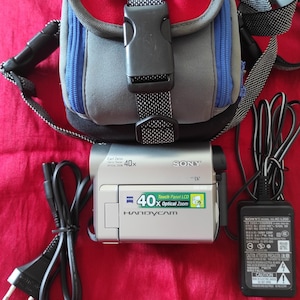 Peut inclure: Caméscope Sony Handycam argenté avec zoom optique 40x, sac pour appareil photo ROWI noir et gris, et adaptateur secteur noir. Le sac est bleu, rouge et gris.