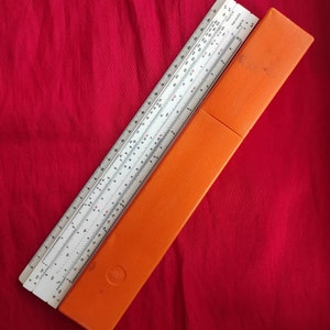 May include: A white slide rule with a black and red scale, in an orange case. The slide rule is a tool used for mathematical calculations.