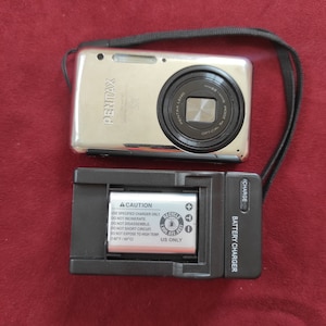May include: A silver digital camera with a black lens and a black strap. The camera is sitting on top of a black battery charger with a white label that says "CAUTION" and "RECYCLE".