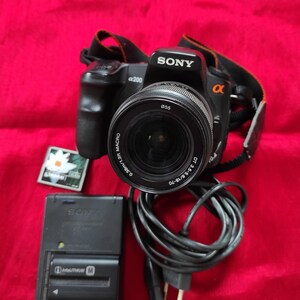 Pode incluir: Câmera digital Sony α200 preta com lente, alça, carregador e cartão pequeno. A lente tem o texto "ø55" e "DT 3.5-5.6/18-70". O carregador tem o texto "InfoLITHIUM M".