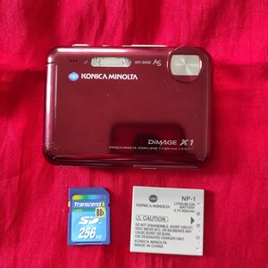 May include: A burgundy Konica Minolta DiMAGE X1 digital camera with an SD card and a battery. The camera has "ANTI-SHAKE As" and "KONICA MINOLTA" printed on it. The SD card is a Transcend 256MB.