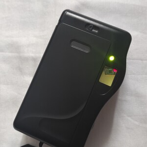 Puede incluir: Una c&aacute;mara compacta negra con un indicador luminoso verde y una correa negra.