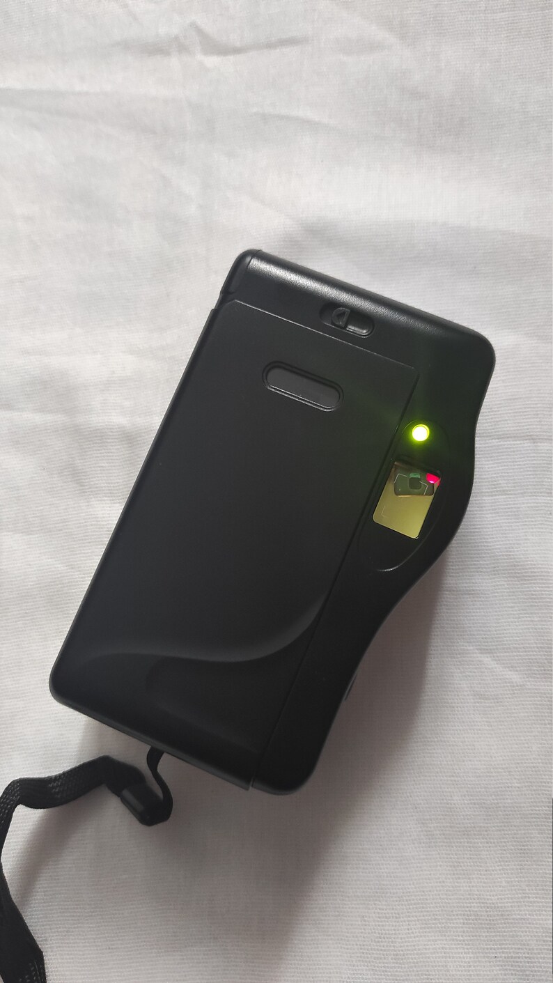 Puede incluir: Una c&aacute;mara compacta negra con un indicador luminoso verde y una correa negra.